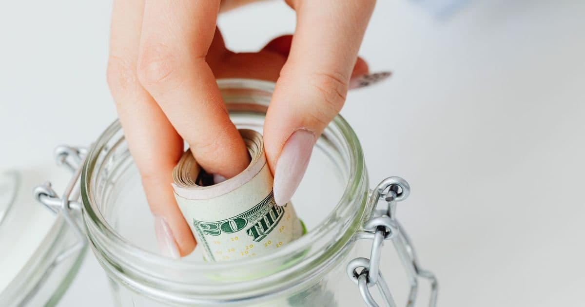 A close-up of a hand placing rolled dollars into a glass jar, symbolizing savings.