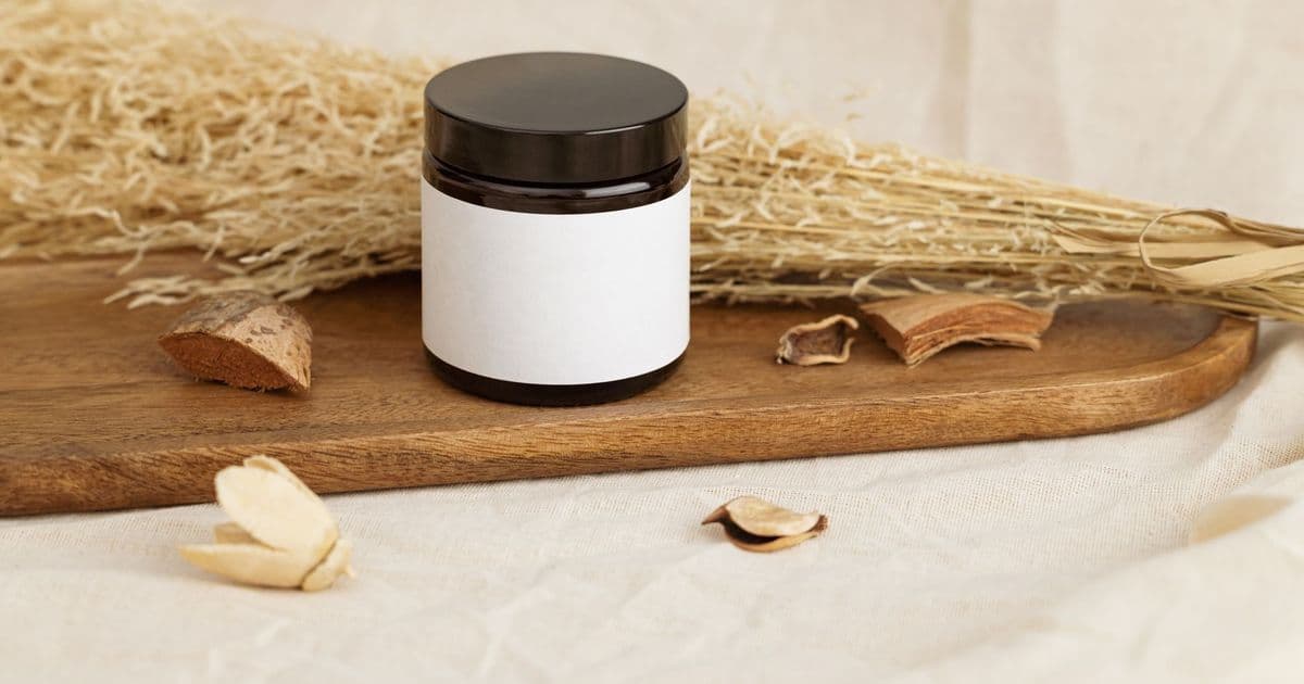Natural cosmetic jar with blank label on a rustic wooden tray surrounded by dried herbs.